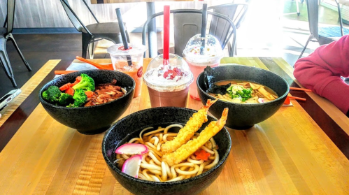 A table with four bowls of noodles and vegetables, accompanied by drinks in clear cups with straws.